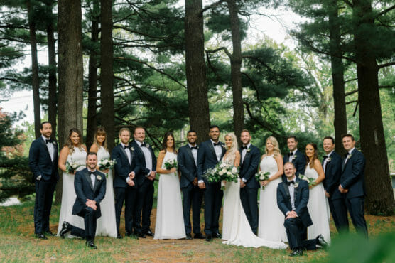 A wedding party of nine women in white dresses and eight men in black tuxedos poses outdoors among tall trees, smiling and holding bouquets, on a grassy, wooded area. by Justin Salem Meyer
