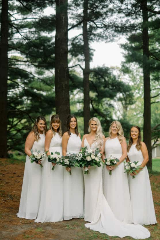 Six women in white dresses stand outdoors in a forested area, holding bouquets of white flowers and greenery. Tall trees surround them, and the group smiles, posing for a formal photo. by Justin Salem Meyer