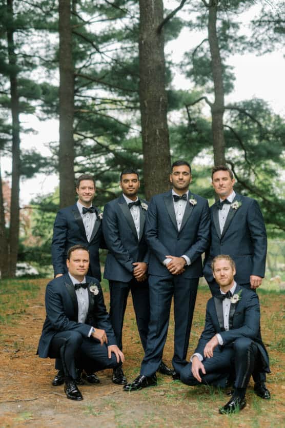 Six men in matching navy tuxedos with bow ties and boutonnieres pose together outdoors in a wooded area, with four standing and two kneeling, surrounded by tall trees and greenery. by Justin Salem Meyer