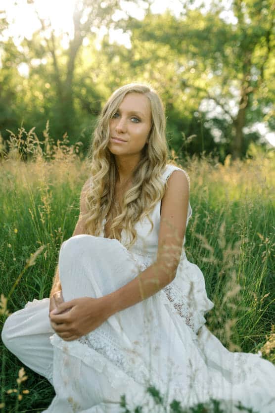A young woman with long blonde hair wearing a white dress sits in a sunlit grassy field, surrounded by tall green grass and trees in the background. by Justin Salem Meyer