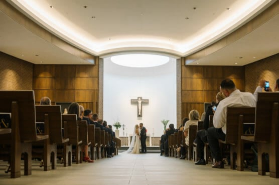 A bride and groom stand at the altar in a modern church, facing each other under a large cross. Guests are seated in rows, some taking photos, watching the wedding ceremony. by Justin Salem Meyer