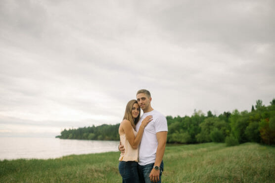 A couple stands close together in a grassy field by the water, with trees in the background and a cloudy sky overhead. The woman rests her hand on the mans chest as they both look at the camera. by Justin Salem Meyer