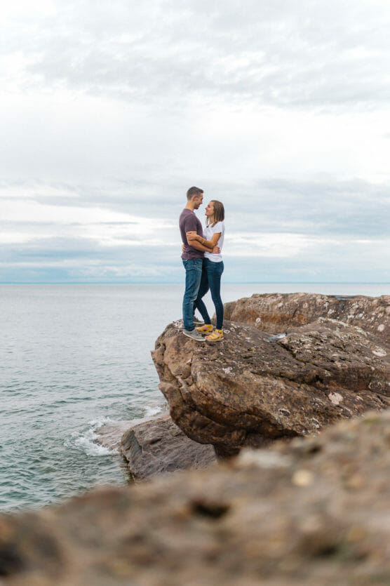 A couple stands close together on a large rocky ledge overlooking the ocean, embracing each other under a cloudy sky. by Justin Salem Meyer