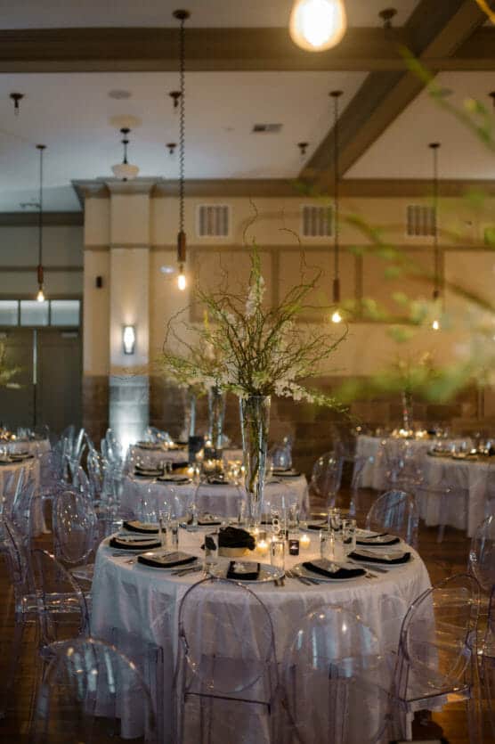 Elegant banquet hall with round tables covered in white cloths, set with black napkins and glassware. Tall floral centerpieces and transparent chairs surround each table. Warm, ambient lighting creates a cozy atmosphere. by Justin Salem Meyer