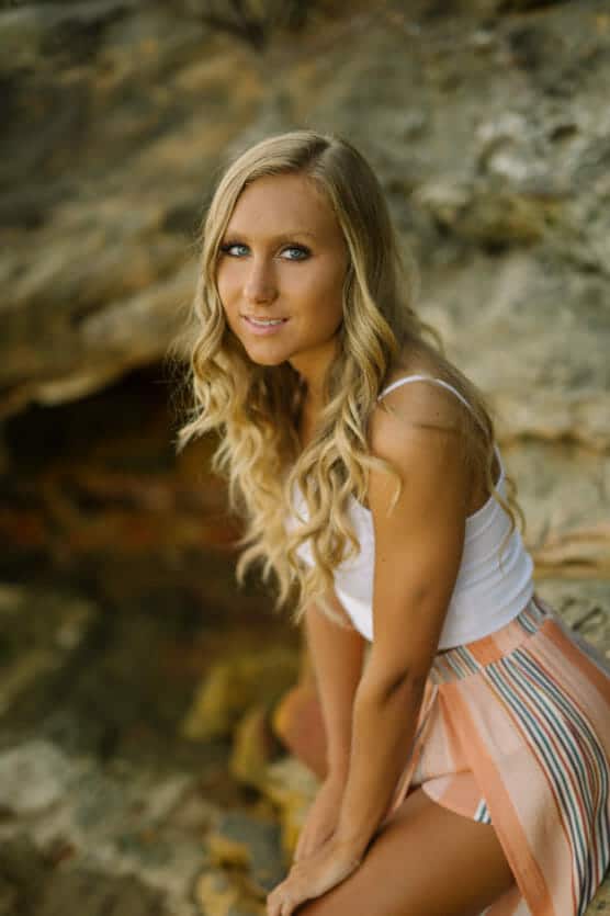A young woman with long, wavy blonde hair and blue eyes is wearing a white tank top and a pastel-colored striped skirt. She is sitting on a rock outdoors and looking at the camera with a gentle expression. by Justin Salem Meyer
