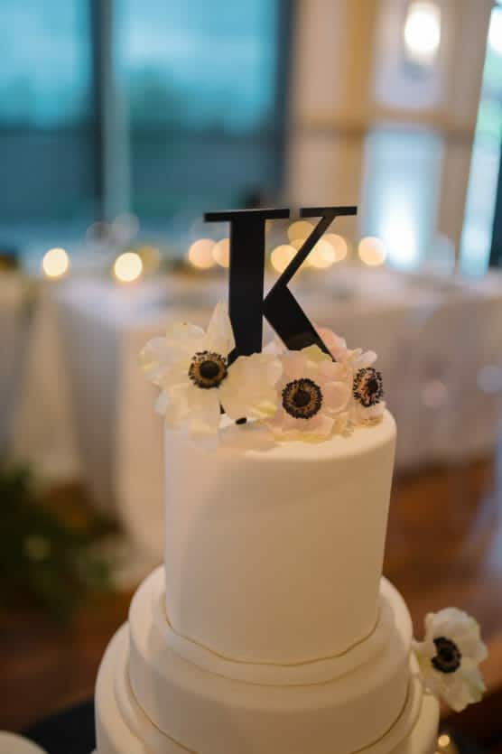 A white tiered cake decorated with white flowers and topped with a large black letter K. The background is softly blurred with lights and a window visible. by Justin Salem Meyer
