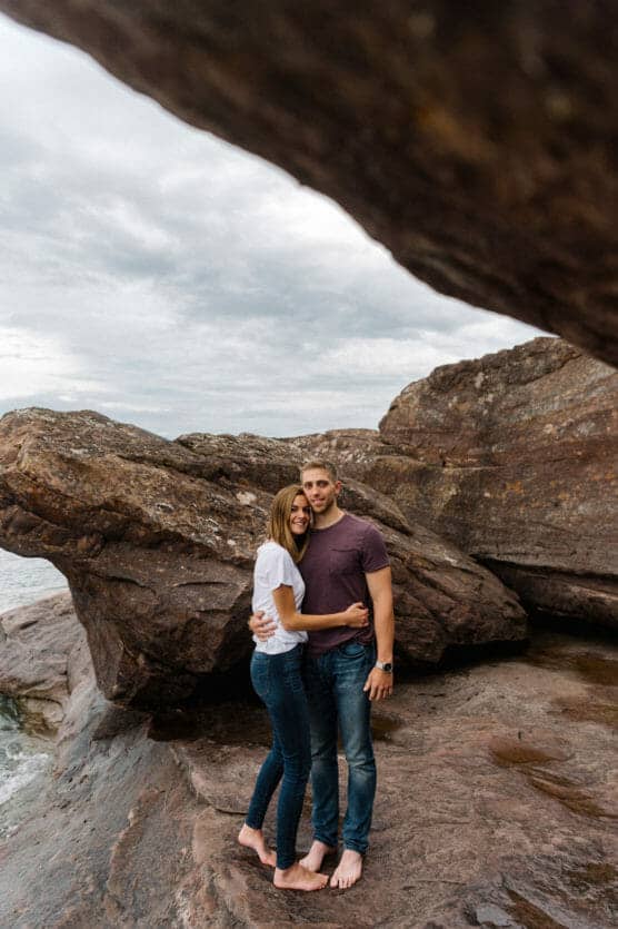 A couple stands barefoot on rocky terrain near a large boulder by the water, embracing and smiling at the camera under a cloudy sky. by Justin Salem Meyer