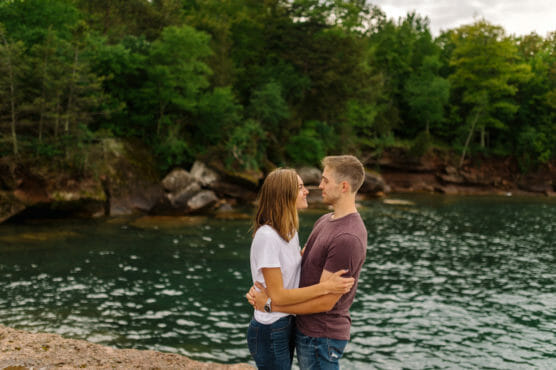 A couple stands embracing and smiling at each other by a lake, surrounded by lush green trees and rocky shoreline. by Justin Salem Meyer