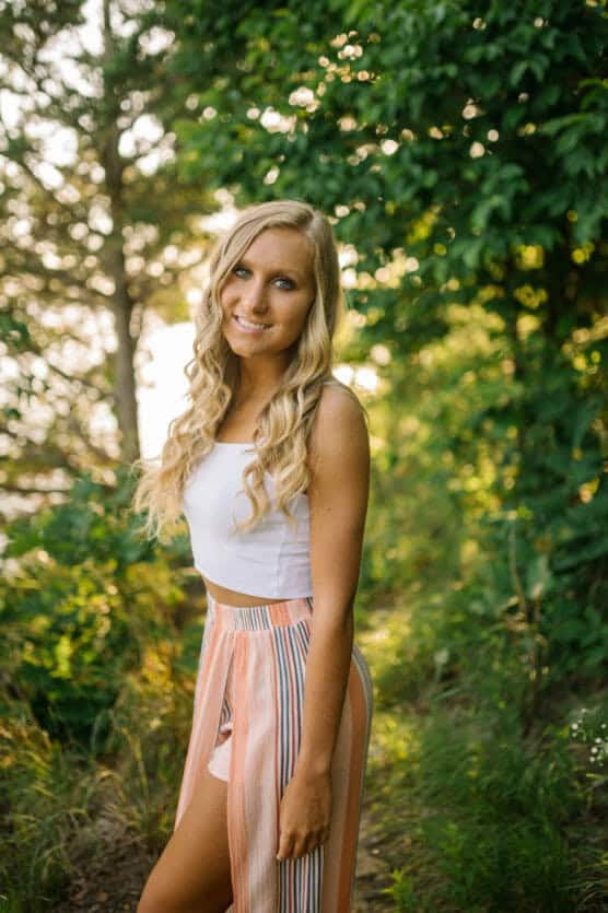 A young woman with long, wavy blonde hair stands outdoors, smiling. She wears a white crop top and peach-striped flowy pants with high slits. Lush green trees and sunlight create a bright, natural background. by Justin Salem Meyer