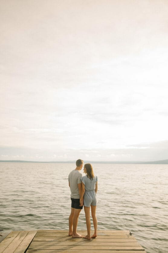 A couple stands barefoot on a wooden dock, holding hands and looking out over a calm lake under a bright, lightly clouded sky. The mood is peaceful and intimate. by Justin Salem Meyer