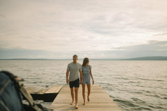 A couple walks barefoot, holding hands on a wooden dock extending into a calm lake, with soft, overcast skies and distant hills in the background. A backpack is visible in the foreground. by Justin Salem Meyer
