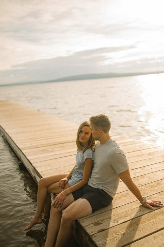 A young couple sits together on a wooden dock by a lake at sunset, with their feet dangling over the water, enjoying the peaceful and warm atmosphere. by Justin Salem Meyer