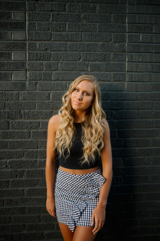 A young woman with long blonde hair and a black sleeveless top stands against a dark brick wall, wearing a black-and-white checkered skirt with a ruffle detail. She looks at the camera with a relaxed expression. by Justin Salem Meyer
