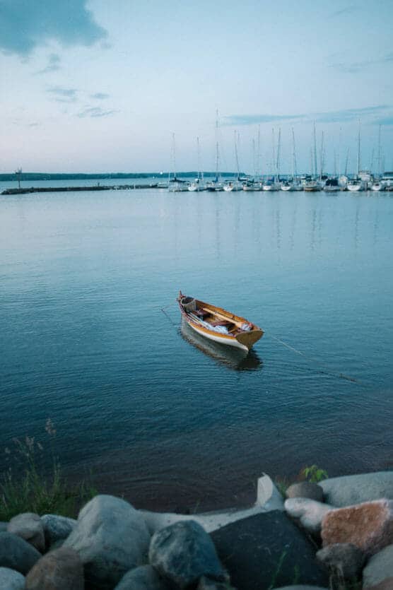A small wooden rowboat floats on calm blue water near the shore, anchored with a rope. In the background, a marina with sailboats is visible under a pale evening sky. Rocks line the waters edge in the foreground. by Justin Salem Meyer