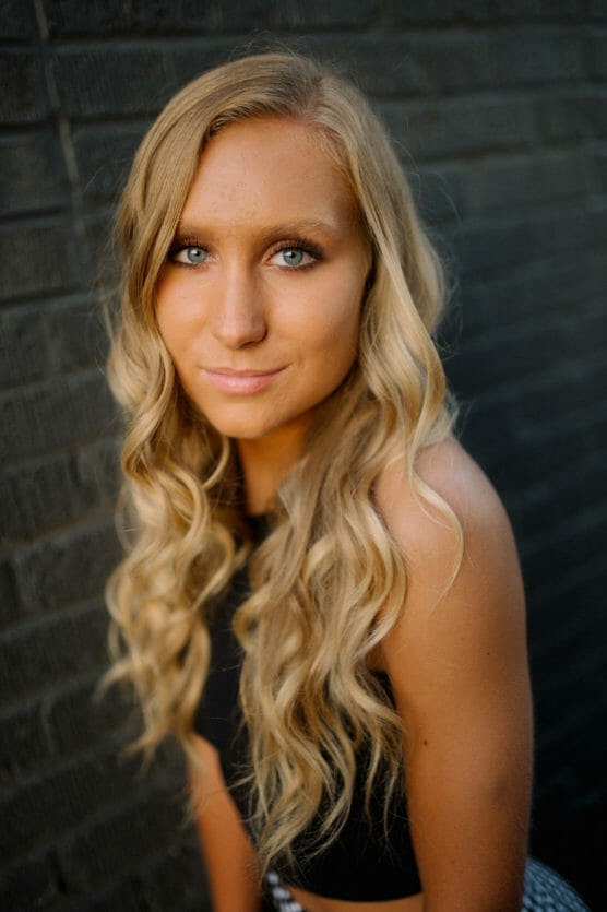 A young woman with long, wavy blonde hair and blue eyes poses against a textured dark brick wall, wearing a black sleeveless top and looking confidently at the camera. by Justin Salem Meyer