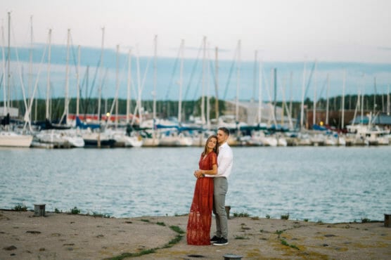A couple stands on a dock embracing, with sailboats and masts lined up in a marina behind them. The woman wears a long red dress; the man is in a white shirt. The water and sky create a calm, romantic backdrop. by Justin Salem Meyer
