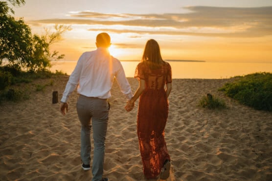 A couple holding hands walks on sandy ground toward a sunset over a calm body of water, surrounded by greenery and a golden sky. by Justin Salem Meyer