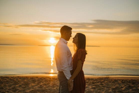 A couple stands close together on a sandy beach at sunset, gazing into each others eyes. The golden sun sets over the calm water, casting a warm, glowing light on the scene. by Justin Salem Meyer