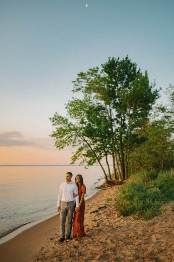A couple stands hand in hand on a sandy beach at sunset, with trees and greenery behind them and calm water beside them under a clear sky with a small moon visible. by Justin Salem Meyer