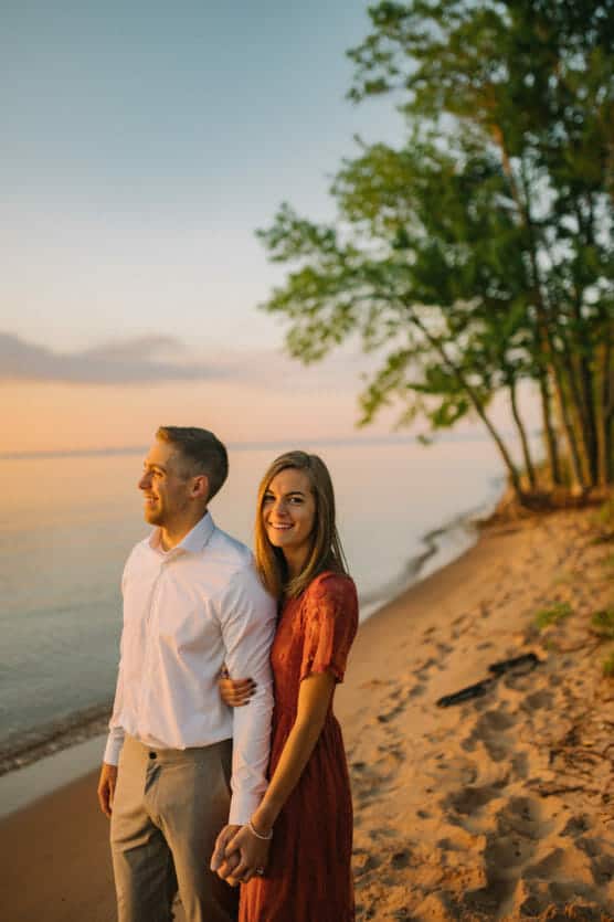 A smiling couple stands hand in hand on a sandy beach at sunset, with calm water and green trees in the background. The man wears a white shirt and gray pants; the woman wears a red dress. by Justin Salem Meyer