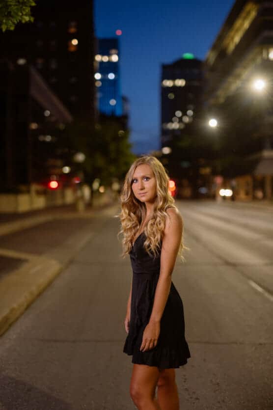 A young woman with long blonde hair wearing a black dress stands in the middle of a city street at dusk, with blurred lights and tall buildings in the background. by Justin Salem Meyer