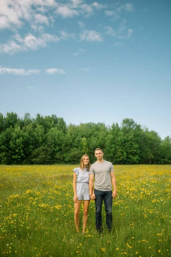 A young couple stands holding hands in a field of yellow wildflowers, with green trees and a blue sky with scattered clouds in the background. Both are smiling and casually dressed. by Justin Salem Meyer