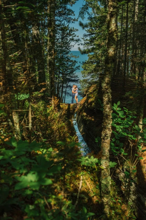 A couple embraces on a narrow wooden bridge in a lush forest, with sunlight filtering through the trees and a glimpse of blue water and sky in the background. by Justin Salem Meyer