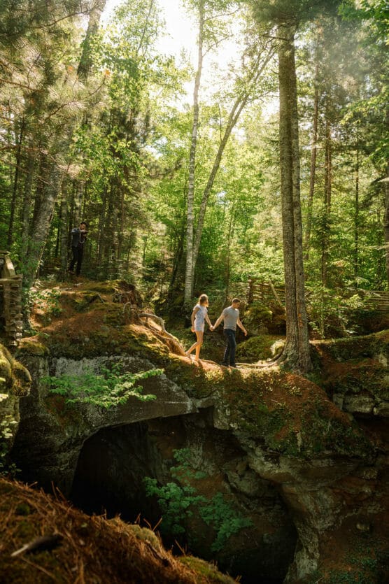 A couple holds hands while walking on a mossy rock bridge in a lush, sunlit forest, with tall trees surrounding them and another person standing on rocks in the background. by Justin Salem Meyer