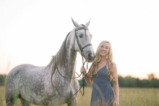 A young woman with long blonde hair in a blue dress stands in a grassy field, smiling, while holding the reins of a gray horse. The background is softly lit, with a blurred treeline at sunset. by Justin Salem Meyer