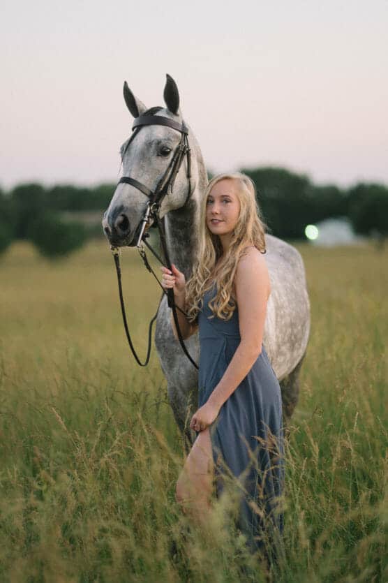 A young woman in a blue dress stands in a grassy field beside a gray horse, holding its reins. The background features blurred trees under a soft evening sky. by Justin Salem Meyer