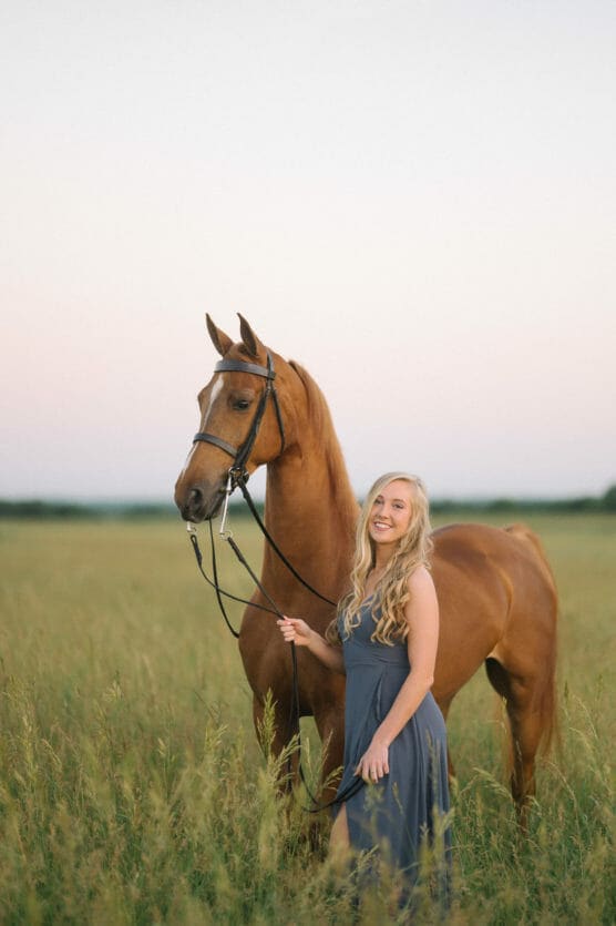 A young woman in a gray dress stands in a grassy field, smiling and holding the reins of a brown horse beside her at sunset. by Justin Salem Meyer