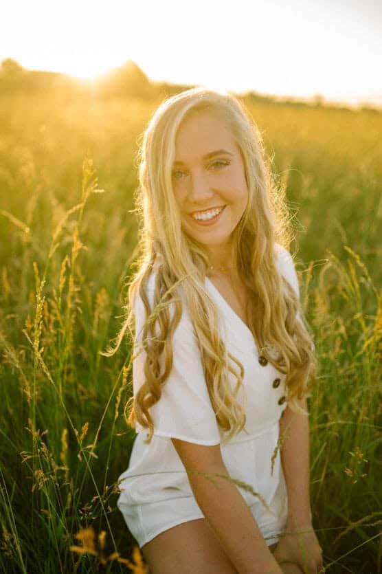 A young woman with long blonde hair and a white buttoned dress sits in tall grass, smiling at the camera with warm sunlight glowing behind her in a field. by Justin Salem Meyer