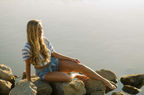 A young woman with long blonde hair sits on rocks beside calm water, wearing a striped crop top and denim shorts, looking thoughtfully into the distance bathed in soft sunlight. by Justin Salem Meyer