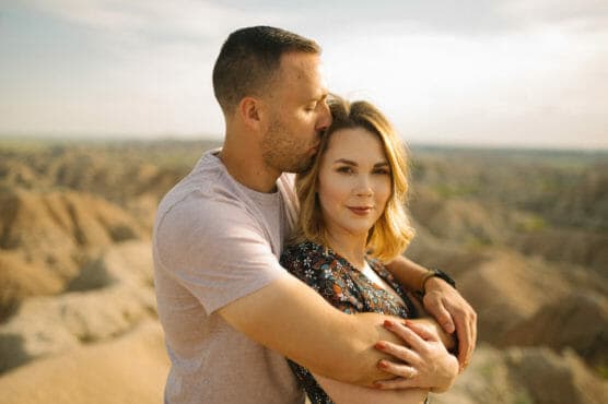A man gently hugs a woman from behind and kisses her on the forehead. They stand outdoors with rocky hills in the background under a bright, sunny sky. The woman faces the camera, softly smiling. by Justin Salem Meyer