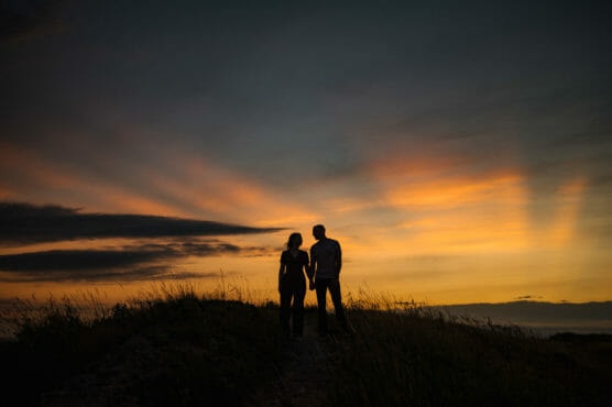 Silhouettes of a couple standing close together on a grassy hill at sunset, with vibrant orange and yellow rays spreading across the sky in the background. by Justin Salem Meyer