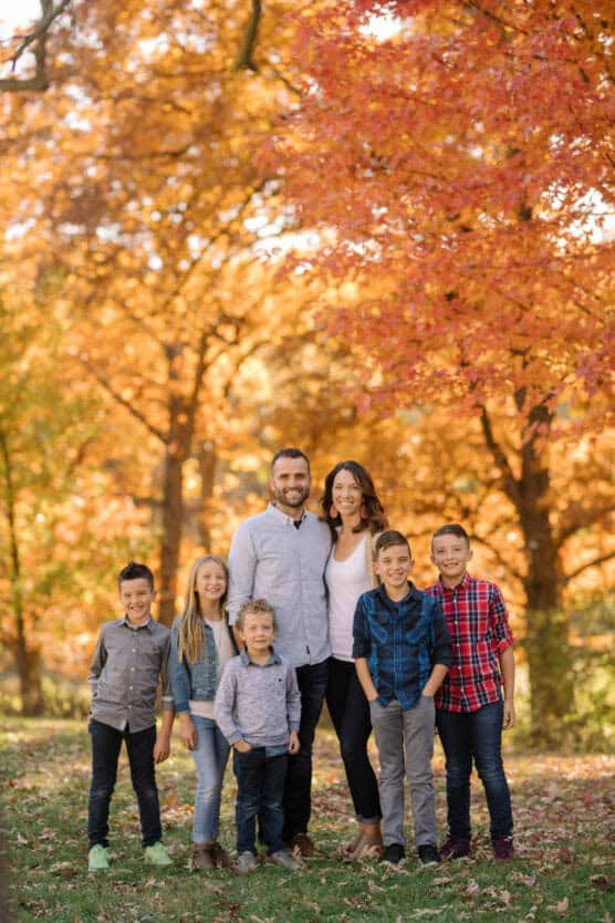 A family of seven—two adults and five children—stands together outside, smiling, with vibrant orange autumn trees in the background. The group is dressed in casual clothes and standing on green grass covered with fallen leaves. by Justin Salem Meyer