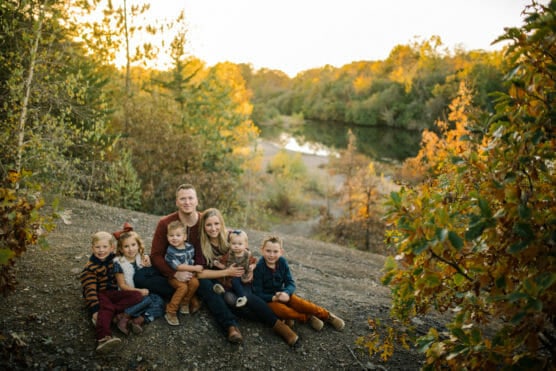 A family of seven, including two adults and five young children, sits together on a rocky slope surrounded by autumn trees. A river and forest are visible in the background under a golden sunset. by Justin Salem Meyer