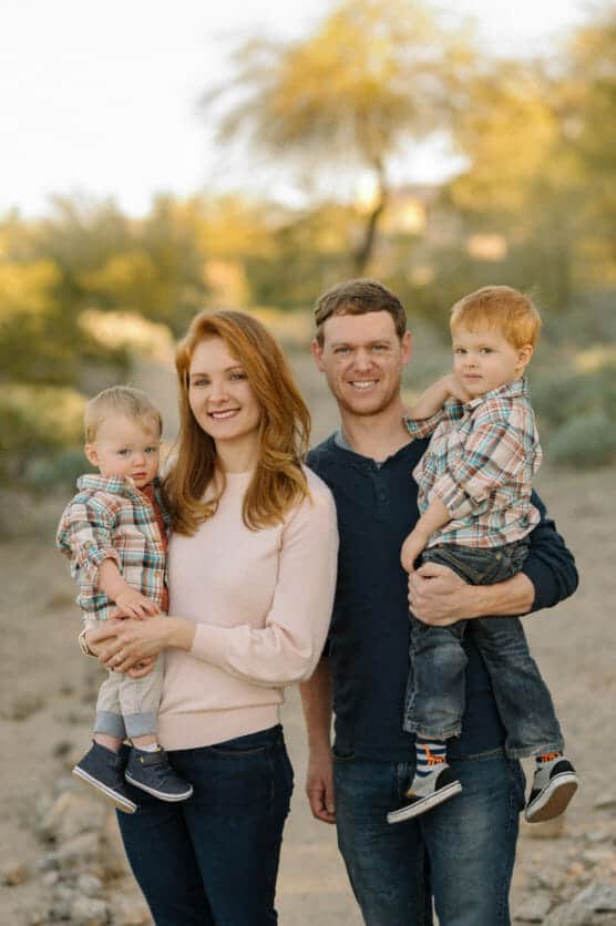 A family of four poses outdoors. The woman holds a young boy, and the man holds another young boy. All are smiling and wearing casual clothes, with trees and soft sunlight in the background. by Justin Salem Meyer