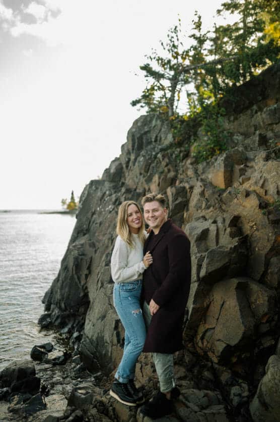 A smiling couple stands on rocky terrain by the water, with trees and a cloudy sky in the background. The woman wears a white sweater and jeans, and the man wears a dark coat. They are close together, looking happy. by Justin Salem Meyer