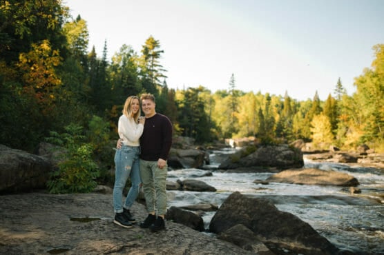 A couple stands together on large rocks beside a flowing river, surrounded by lush green trees under a clear blue sky in a forested area. by Justin Salem Meyer