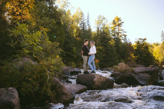 A couple stands on large rocks in the middle of a flowing river, surrounded by lush green trees and forest under a clear sky, gazing at each other. by Justin Salem Meyer