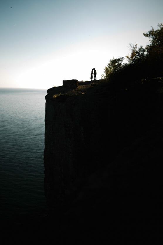 A couple stands close together on the edge of a high cliff overlooking a calm body of water at sunset, silhouetted against the sky with trees nearby. by Justin Salem Meyer