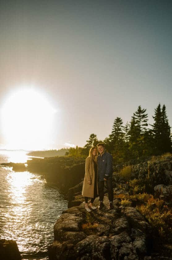 A couple stands on rocky terrain by the water at sunset, with trees in the background and sunlight reflecting on the water. by Justin Salem Meyer