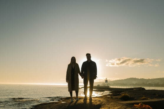 A couple holding hands stands on a rocky shore at sunset, with the ocean, a distant pier, and a lighthouse in the background. The sun is low in the sky, casting a warm glow and creating their silhouettes. by Justin Salem Meyer