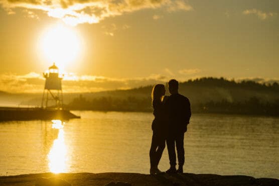 A couple stands together near the water, silhouetted against a golden sunset with a lighthouse and distant hills in the background. by Justin Salem Meyer