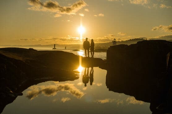 Two people stand on rocks by a calm body of water at sunset, their silhouettes reflected in the water. The sun is low in the sky, creating a golden glow, with a lighthouse and distant hills in the background. by Justin Salem Meyer