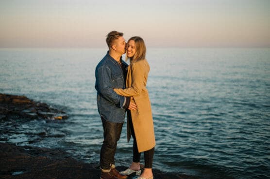 A couple stands on a rocky shore by the sea at sunset. The man, in a denim jacket, kisses the woman, who wears a beige coat, on the forehead. They both smile and embrace, with calm water and a soft pastel sky behind them. by Justin Salem Meyer