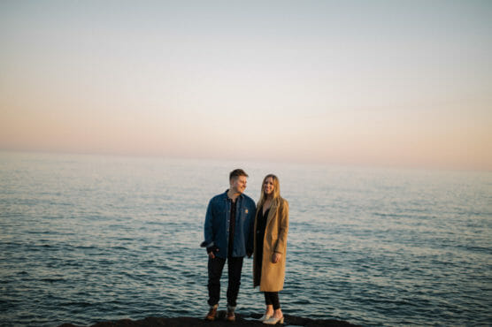 Two people stand on a rocky shore in front of a calm sea at sunset. One wears a denim jacket, the other a tan coat. The sky is soft pastel colors, and both are smiling gently at the camera. by Justin Salem Meyer