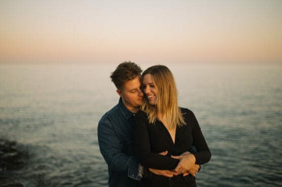 A couple embraces by the water at sunset. The man in a denim shirt hugs the woman from behind; she smiles and looks to the side. The soft light reflects off the calm sea in the background. by Justin Salem Meyer