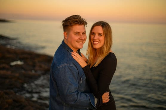 A smiling couple embraces by the shoreline at sunset, with calm water and a warm orange sky in the background. The man wears a denim jacket, and the woman wears a black top. by Justin Salem Meyer